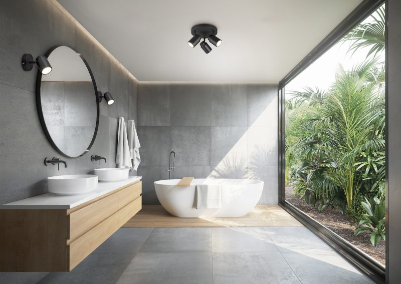 Grey concrete tiled bathroom with opening to a jungle garden a round mirror and a bathtub