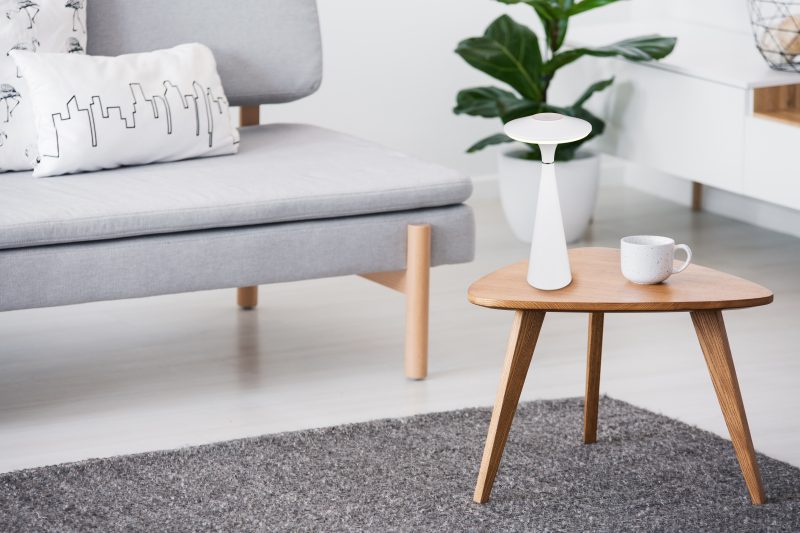 Cup on a wooden coffee table and blurry foreground with graphic pillows on a gray sofa in a white living room interior